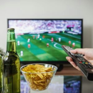 A person holding a remote while watching soccer on TV with beer and snacks on the table.